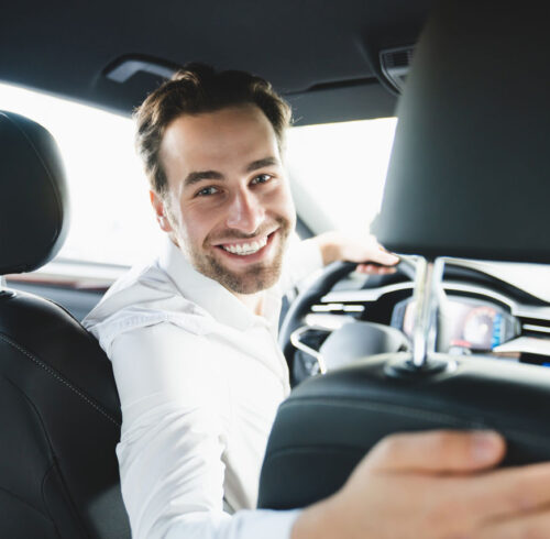Taxi driver smiling while turning over to the backseat looking talking with clients. Successful businessman husband looking at the backseat at dealer shop before buying a car