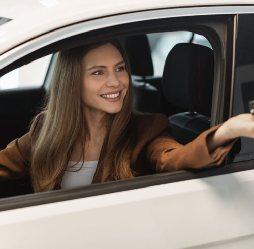 Happy young female customer sitting inside new car, taking auto key from salesman at modern dealership store, panorama. Cheerful millennial woman buying or renting automobile at showroom