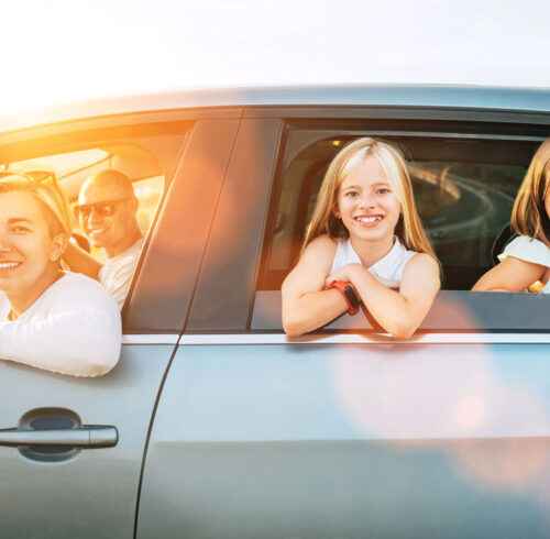 Happy young couple with two daughters inside car during auto trop. They are looking out auto windows, smiling, laughing during road trip. Family values, traveling concept side car view photo.